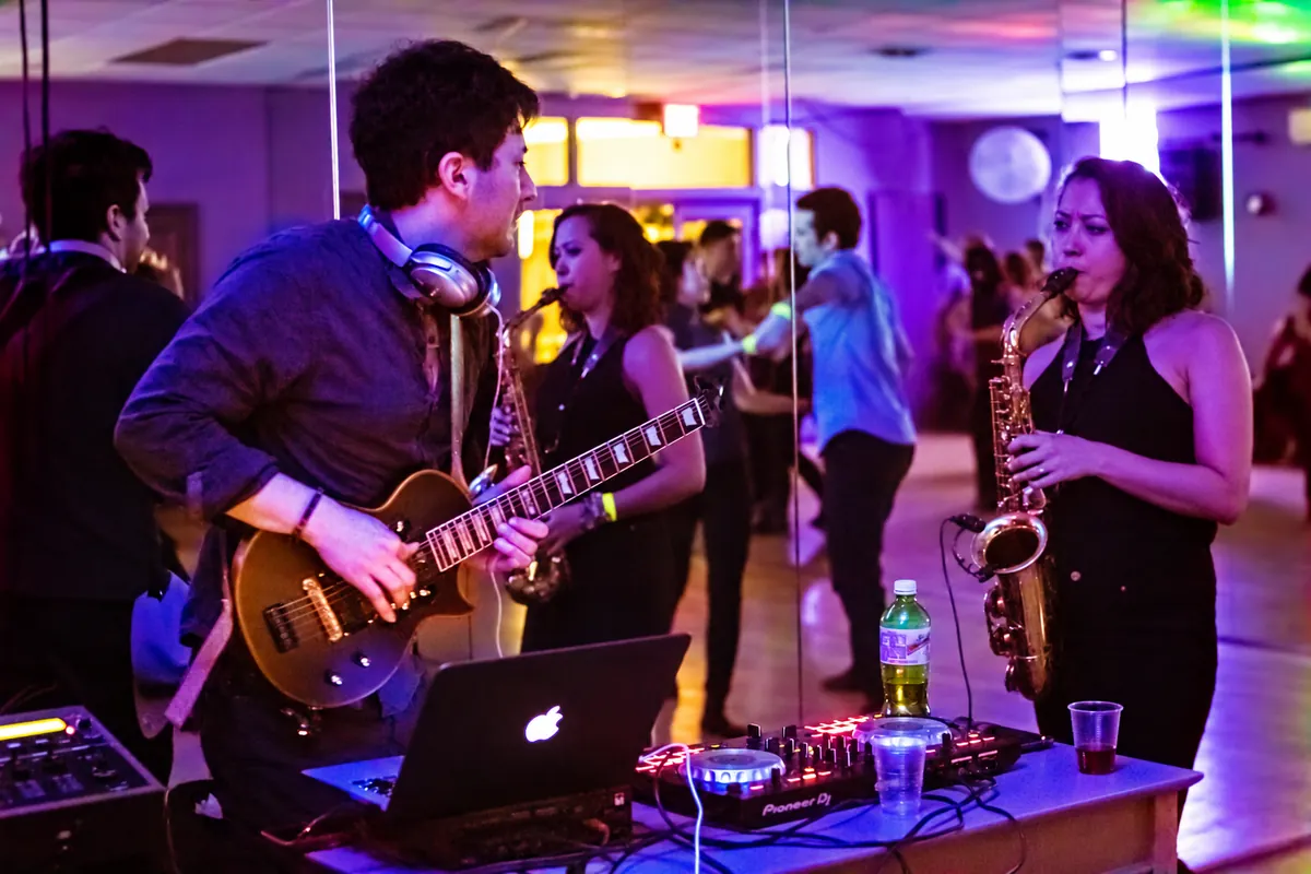Sasha playing electric guitar alongside a saxophonist, with the DJ equipment in front of them